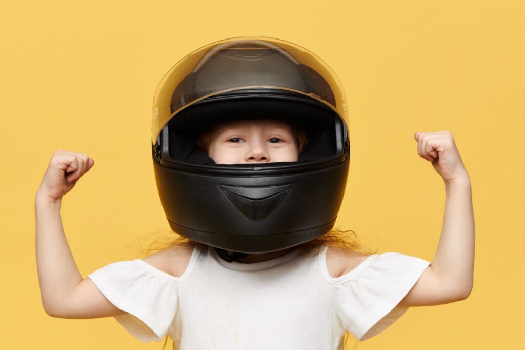 Kleines Mädchen posiert vor gelber Wand, trägt schwarzen Sicherheitsmotorradhelm